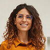A woman with curly brown hair and glasses, wearing an orange shirt, smiles while standing in front of a plain light background.
