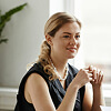 A woman with blonde hair tied back, wearing a sleeveless black top and pearl necklace, sits indoors near a window, holding her hands together and smiling.