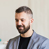 A man with a short beard and short hair, wearing a gray blazer over a black shirt, is looking slightly down and smiling indoors.