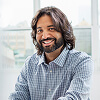 A man with long hair and a beard, wearing a checked shirt, sits at a desk and smiles at the camera in a bright indoor setting.