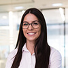 A woman with long dark hair and glasses, wearing a white shirt, smiles while standing in a modern office setting.