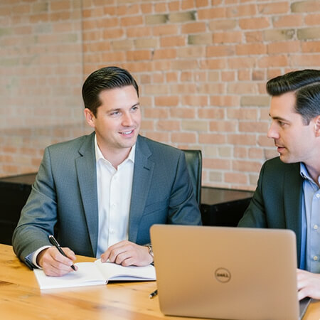 Two men in business attire sit at a wooden table, one taking notes in a notebook while the other uses a laptop, in an office with a brick wall background.