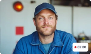 A man in a blue cap and jacket looks at the camera. A red light and timer reading 02:20 appear in the background.