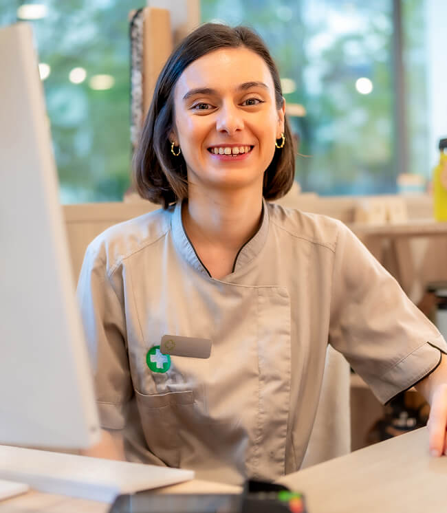 A woman in a light gray uniform sits at a desk in a pharmacy, smiling at the camera with a computer monitor in front of her.