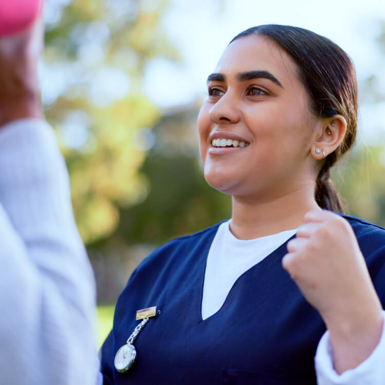 A woman in navy medical scrubs and a white undershirt smiles outdoors, looking at a person holding a pink object. Trees are visible in the background.