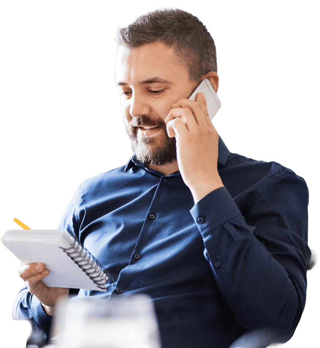 Man in a blue shirt talking on a smartphone while holding a notepad and pencil.
